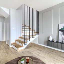 Scandywork sp z o o - Minimalistyczne wnętrze z drewnianymi schodami o czarnych pionowych balustradach i białych podstopnicach. Szare ściany z dekoracyjnymi listwami, podłoga z płytek imitujących jasne drewno. Duży...