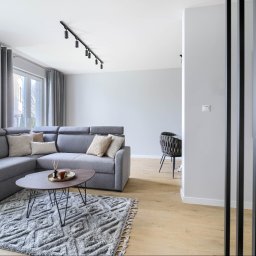 Scandywork sp z o o - Nowoczesny salon z szarą narożną sofą, okrągłym stolikiem kawowym na wzorzystym dywanie i jasną drewnianą podłogą. Czarne oświetlenie szynowe i dekoracyjne lamele dodają charakteru.