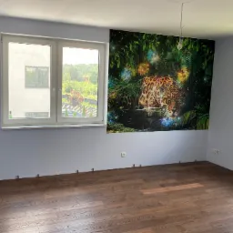 Pokój z nową, ciemną podłogą z paneli i świeżo pomalowanymi na szaro ścianami. Na jednej ze ścian fototapeta z motywem jaguara w dżungli. Widoczne gniazdka elektryczne i brak listew przypodłogowych.