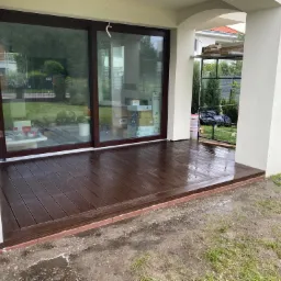 Nowo wybudowany taras z kompozytowych desek w kolorze ciemnego brązu, z czerwoną obwódką z cegieł, przylegający do budynku z dużymi przesuwnymi drzwiami i białymi kolumnami, otoczony mokrą ziemią...