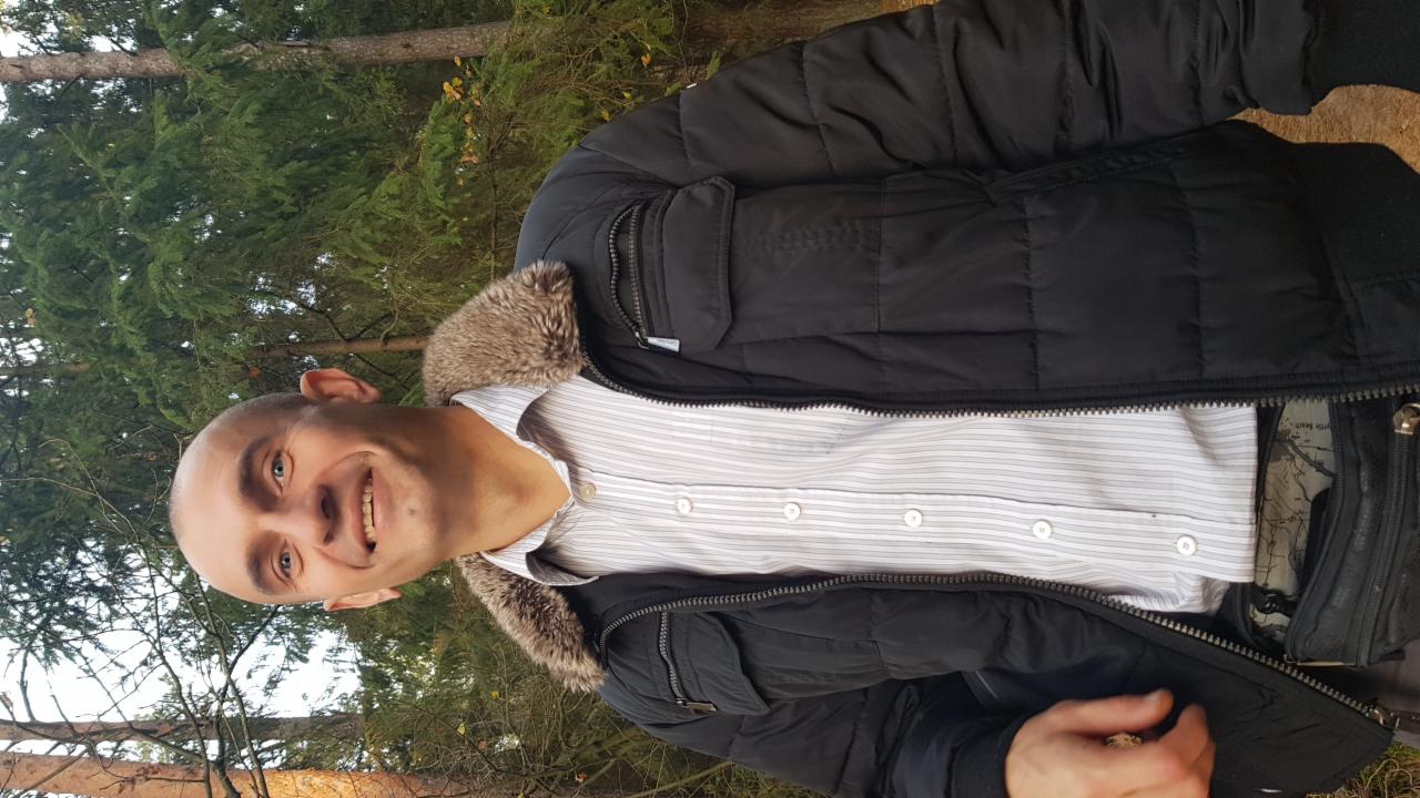 Uśmiechnięty mężczyzna w czarnej kurtce z futrzanym kołnierzem na tle lasu. Widoczne drzewa iglaste i liściaste, naturalne światło.