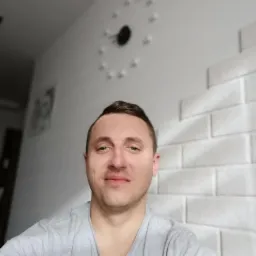 Portret mężczyzny na tle białej ściany z dekoracyjnym zegarem i imitacją cegły, wykonany z bliskiej odległości.