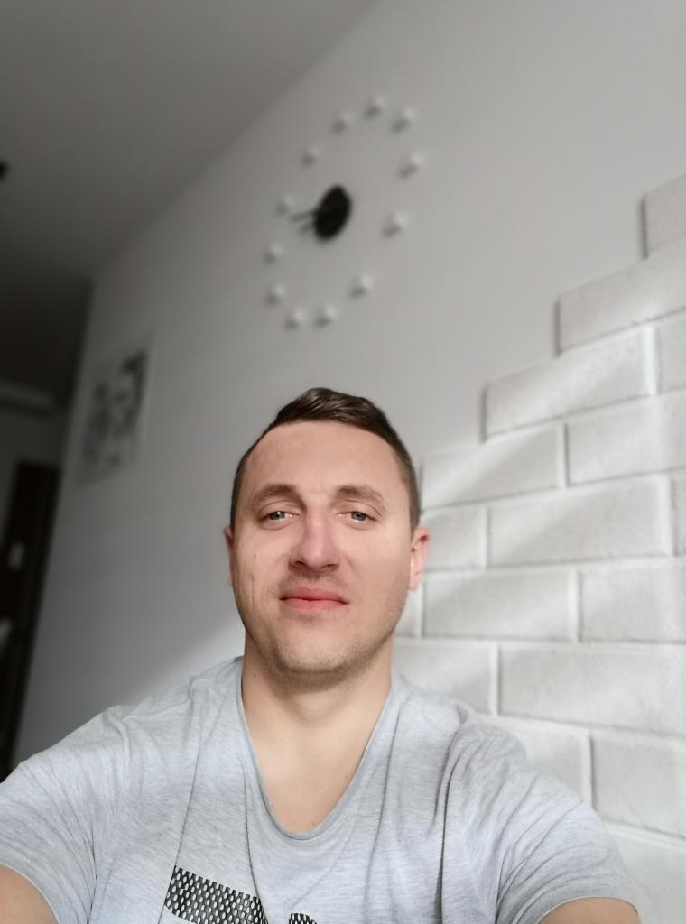 Portret mężczyzny na tle białej ściany z dekoracyjnym zegarem i imitacją cegły, wykonany z bliskiej odległości.