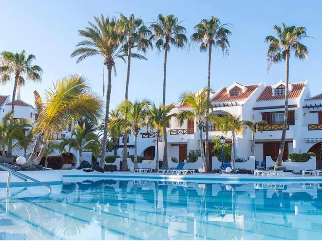 Kompleks apartamentów z basenem i palmami w słoneczny dzień. Białe budynki z brązowymi okiennicami i czerwonymi dachówkami odbijają się w lustrzanej powierzchni basenu.