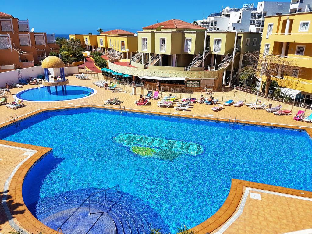 Kompleks basenowy z mozaiką w kształcie napisu, otoczony leżakami i budynkami w słoneczny dzień.