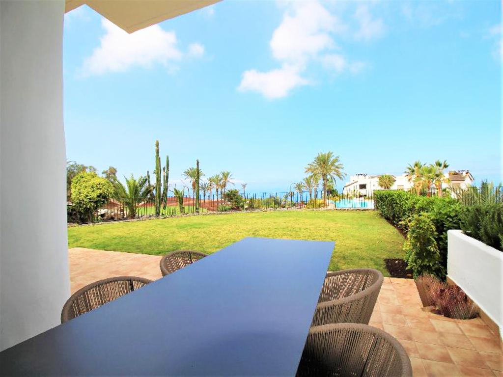 Widok z tarasu na zadbany trawnik z palmami i kaktusami, w tle budynek z basenem i błękitne niebo, na pierwszym planie stół z krzesłami.