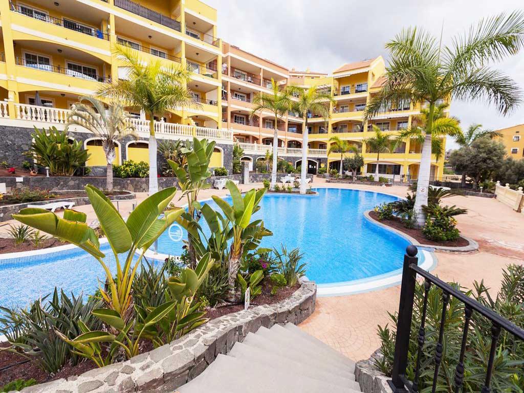 Kompleks apartamentowy z żółtą elewacją i balkonami, otoczony palmami i basenem o nieregularnym kształcie, widok z góry schodów.