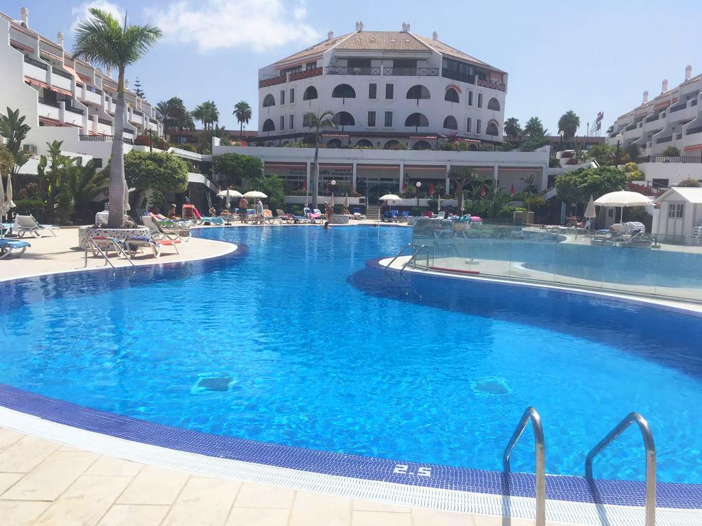 Duży, błękitny basen w kompleksie hotelowym z palmami i białymi budynkami w tle, widok w słoneczny dzień.