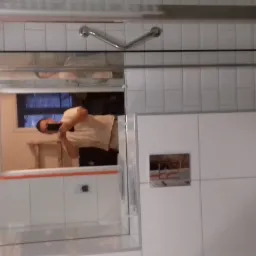 Nowoczesna łazienka z białymi płytkami, kabiną prysznicową ze szklanymi drzwiami i wiszącą toaletą. W lustrze odbija się osoba robiąca zdjęcie telefonem oraz fragment drabiny.