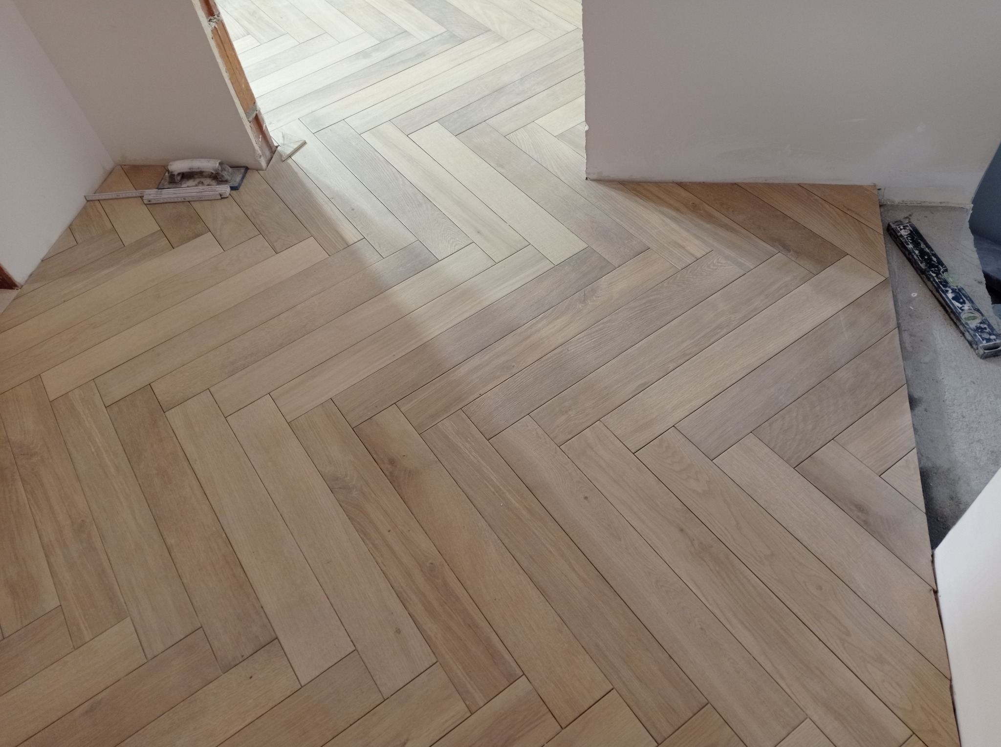 Ułożona podłoga z jasnych paneli w jodełkę francuską, widoczne narzędzia glazurnicze przy ścianie. Częściowo wykończone pomieszczenie.