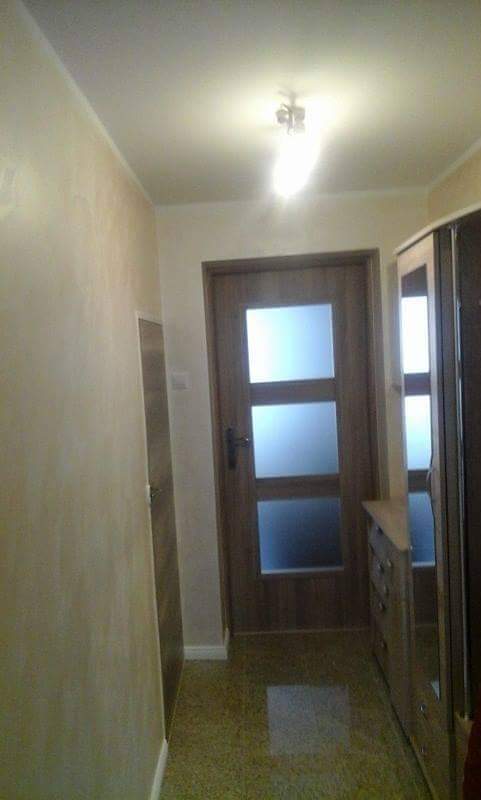 Korytarz po odświeżeniu z drewnianymi drzwiami z trzema szybami i szafą z szufladami w kolorze drewna, widoczna struktura malowania ścian. Na podłodze widoczna mozaika.