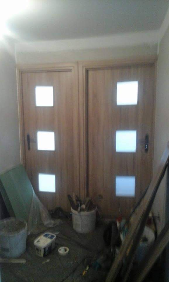 Dwa drewniane drzwi wewnętrzne z trzema kwadratowymi przeszkleniami, widoczne narzędzia i materiały malarskie na podłodze, w trakcie prac wykończeniowych.