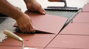 Układanie brązowych płytek podłogowych, widoczna kielnia z drewnianą rączką i rozprowadzony klej na podłożu. Płytka trzymana w dłoniach, gotowa do ułożenia.