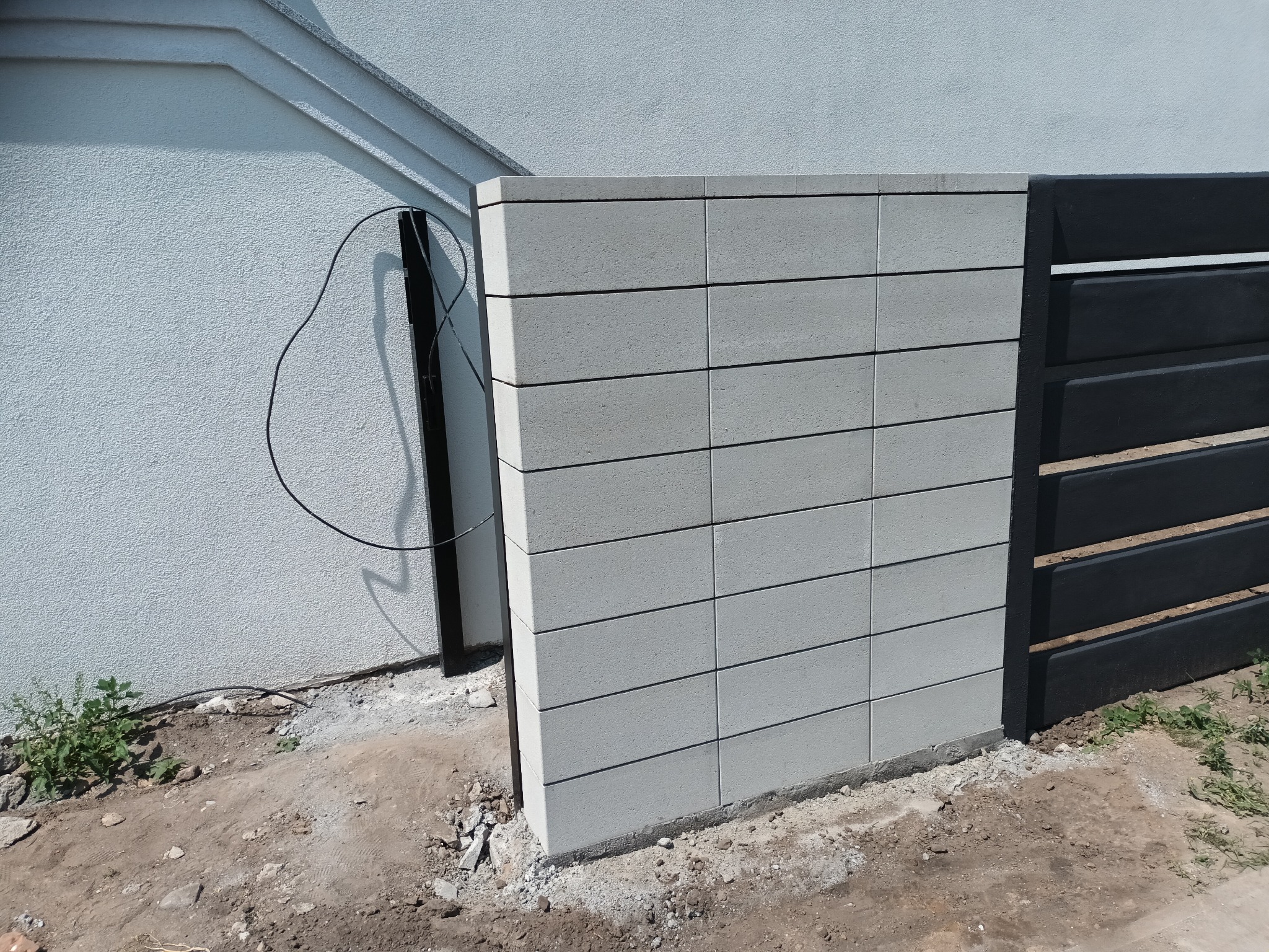 Fragment nowo postawionego ogrodzenia: szary, betonowy słupek z poziomych bloczków sąsiadujący z czarnym, poziomym panelem, widoczne ślady po budowie na ziemi.