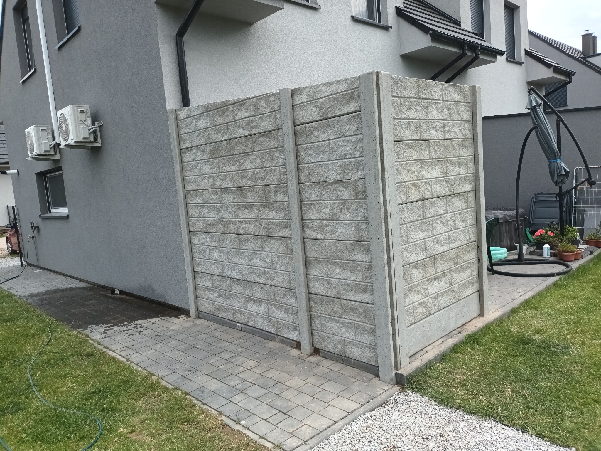 Nowoczesny, szary, dwusegmentowy płot z prefabrykatów betonowych o fakturze cegły, oddzielający posesję od sąsiada, widok z boku na tle domu z klimatyzatorami.