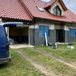 Dom jednorodzinny w trakcie ocieplania elewacji styropianem, okna zabezpieczone folią, widoczny otwarty garaż z materiałami budowlanymi i samochód dostawczy z otwartą klapą bagażnika.