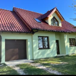 Nowo wybudowany, parterowy dom z poddaszem użytkowym, pokryty czerwoną dachówką, z oknem dachowym i garażem w bryle budynku, pomalowany na jasnozielony kolor.