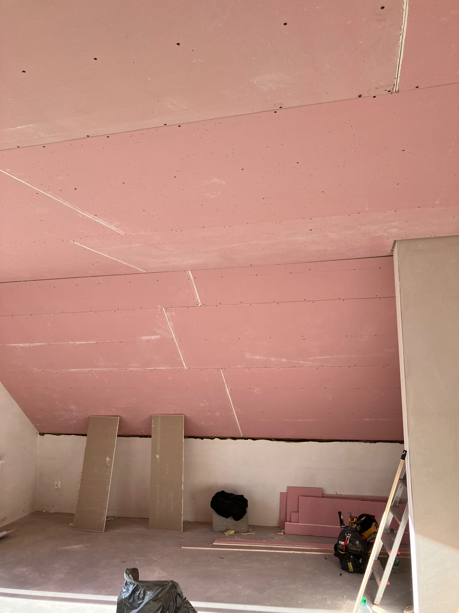 Poddasze w trakcie prac wykończeniowych: zamontowane płyty gipsowo-kartonowe w kolorze różowym na skosach, widoczne łączenia płyt, narzędzia pracy, drabina oparta o ścianę, worki na śmieci.