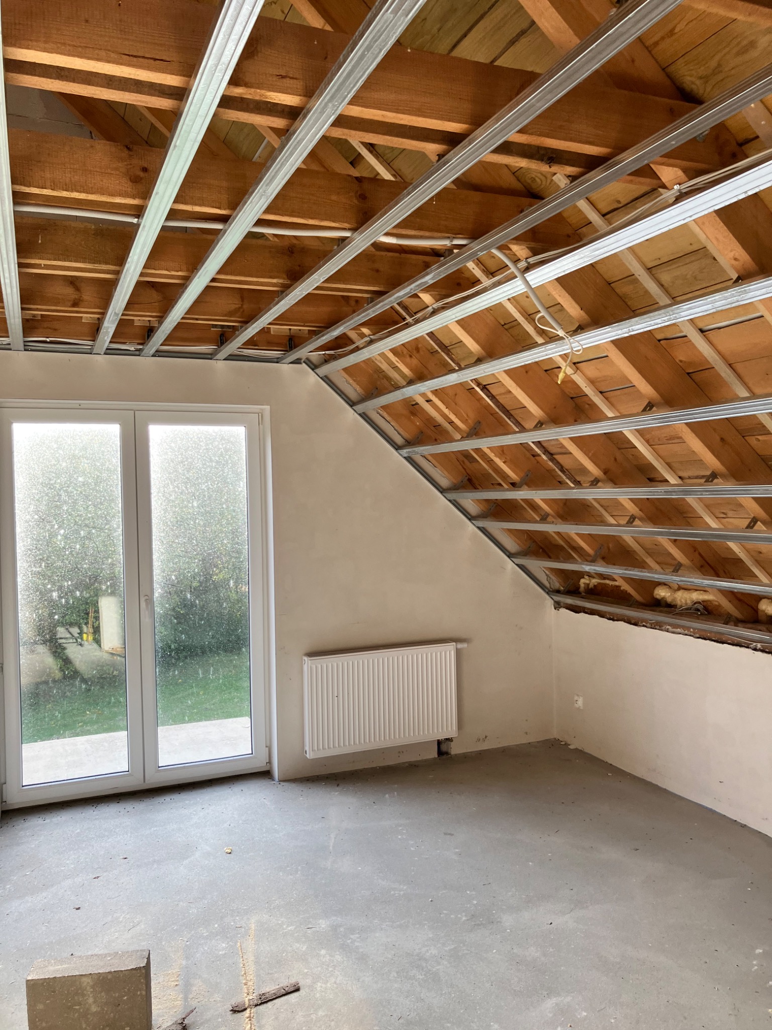 Poddasze w Pozezdrzu w trakcie adaptacji, widoczne drewniane krokwie, metalowa konstrukcja pod płyty gipsowe, okno balkonowe i grzejnik na ścianie.