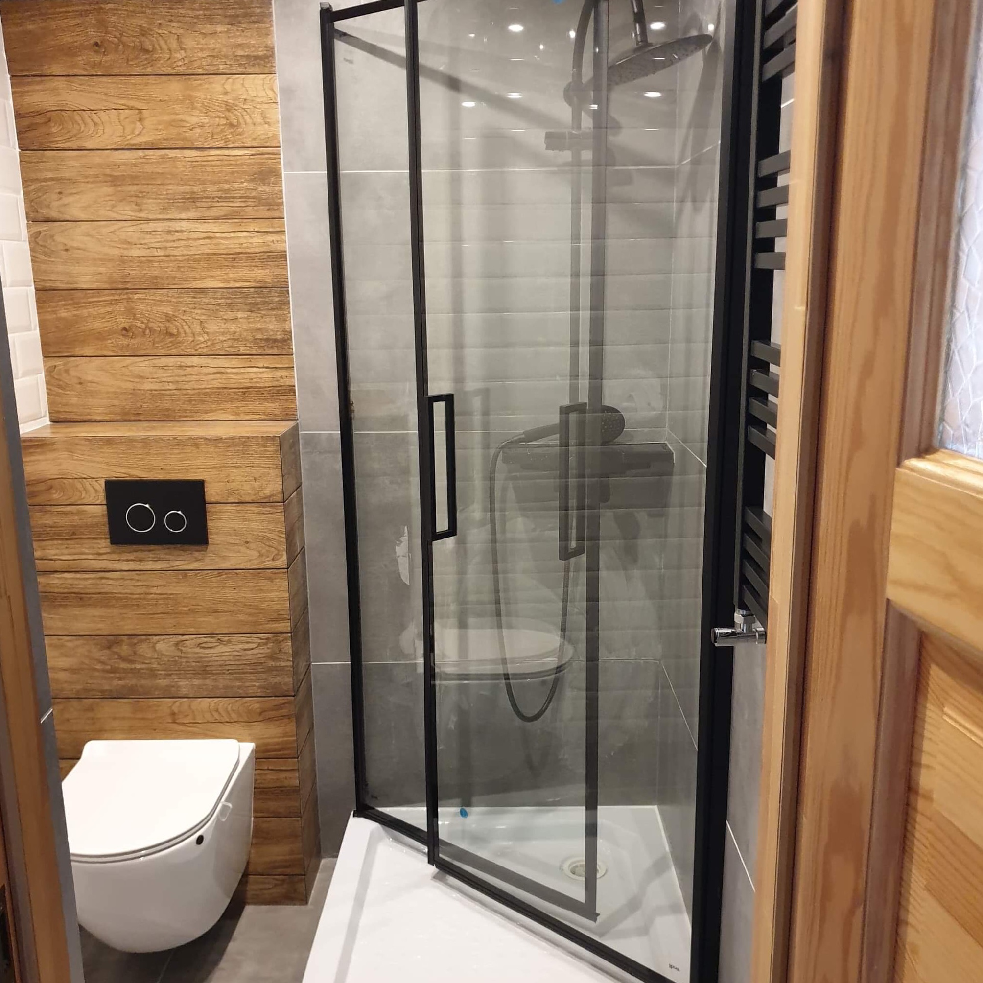 Nowoczesna łazienka z kabiną prysznicową w czarnej ramie, drewnianą boazerią i podwieszaną toaletą z czarnym panelem sterującym.