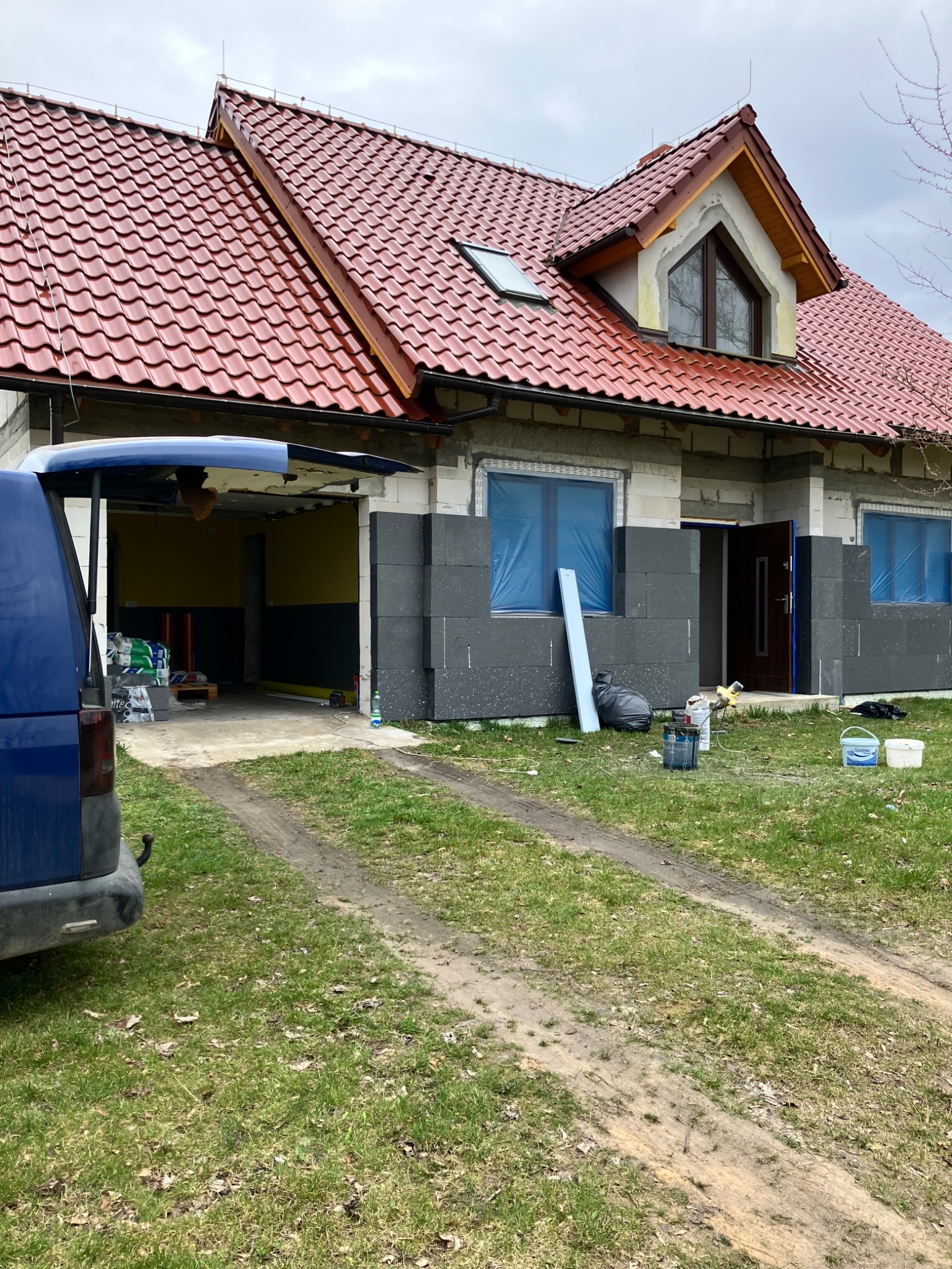 Dom jednorodzinny w trakcie ocieplania elewacji styropianem, okna zabezpieczone folią, widoczny otwarty garaż z materiałami budowlanymi i samochód dostawczy z otwartą klapą bagażnika.