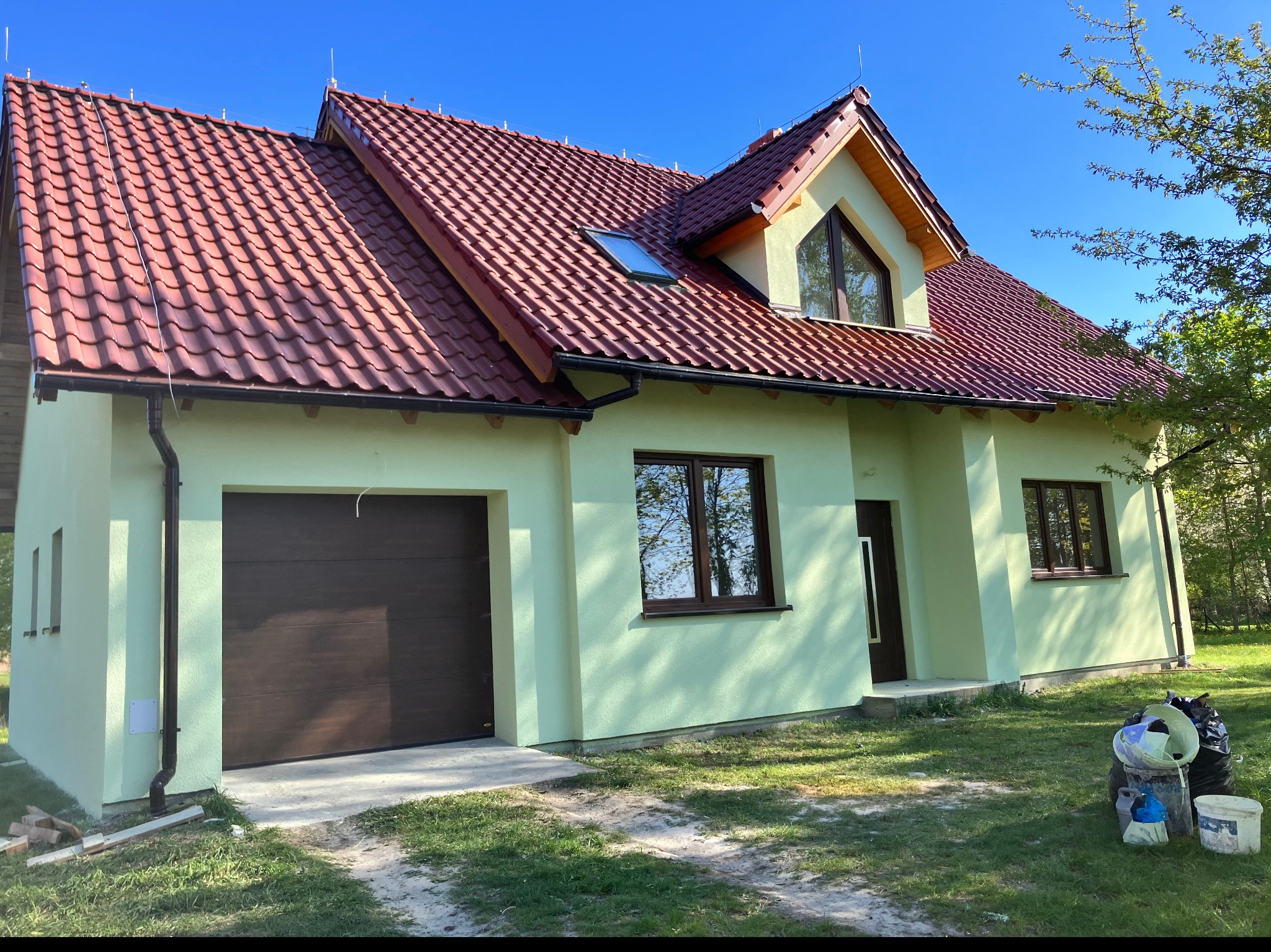 Nowo wybudowany, parterowy dom z poddaszem użytkowym, pokryty czerwoną dachówką, z oknem dachowym i garażem w bryle budynku, pomalowany na jasnozielony kolor.