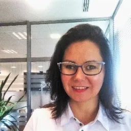 Uśmiechnięta kobieta w okularach w biurowym otoczeniu z rośliną doniczkową i szklanymi ściankami działowymi.