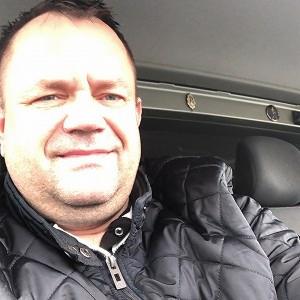 Uśmiechnięty mężczyzna w czarnej kurtce siedzi w fotelu busa, patrząc w obiektyw. Widoczne wnętrze pojazdu, ciemne obicia foteli i fragment deski rozdzielczej.