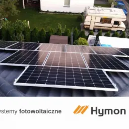 Panele słoneczne na dachu domu jednorodzinnego z widocznym kamperem i zadbanym ogrodem w tle, logo firmy Hymon w dolnej części zdjęcia.