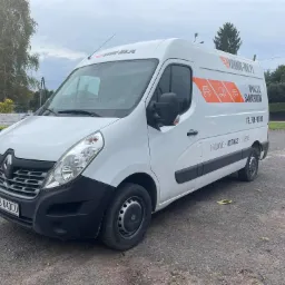 Biały Renault Master z pomarańczowym logo firmy Dynamic-MK.pl oferującej wynajem samochodów, zaparkowany na żwirowym parkingu w pochmurny dzień.
