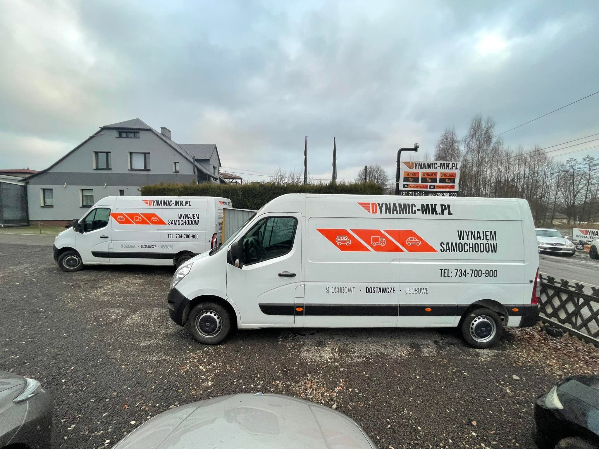 Dwa białe busy z pomarańczowymi grafikami i napisem 'DYNAMIC-MK.PL WYNAJEM SAMOCHODÓW TEL: 734-700-900 9-OSOBOWE DOSTAWCZE OSOBOWE' stojące na parkingu przed budynkiem w pochmurny dzień.