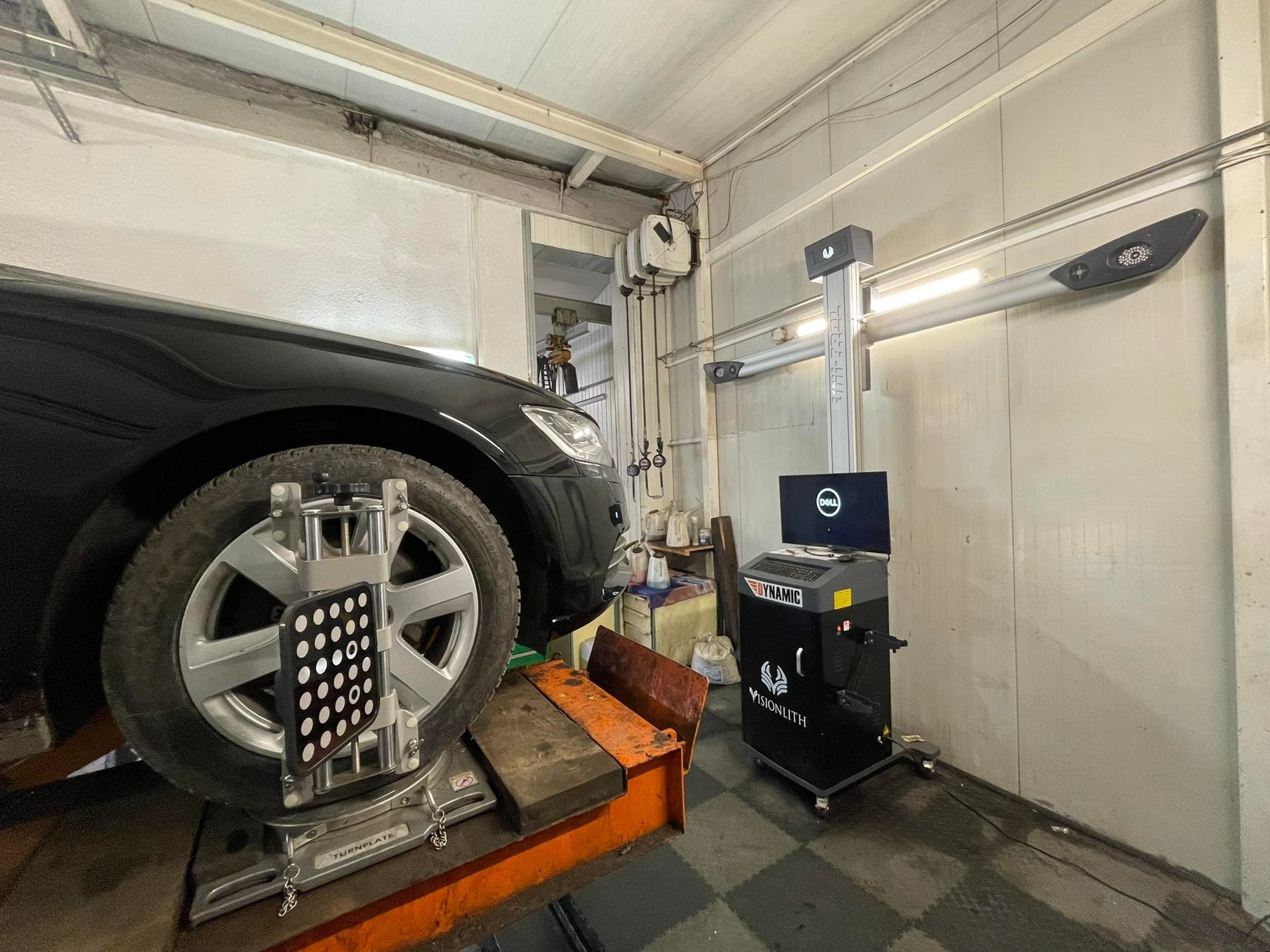 Czarny samochód na stanowisku do geometrii kół z zamontowanymi tarczami pomiarowymi, widoczny komputer z oprogramowaniem, warsztatowy sprzęt do ustawiania zbieżności kół.