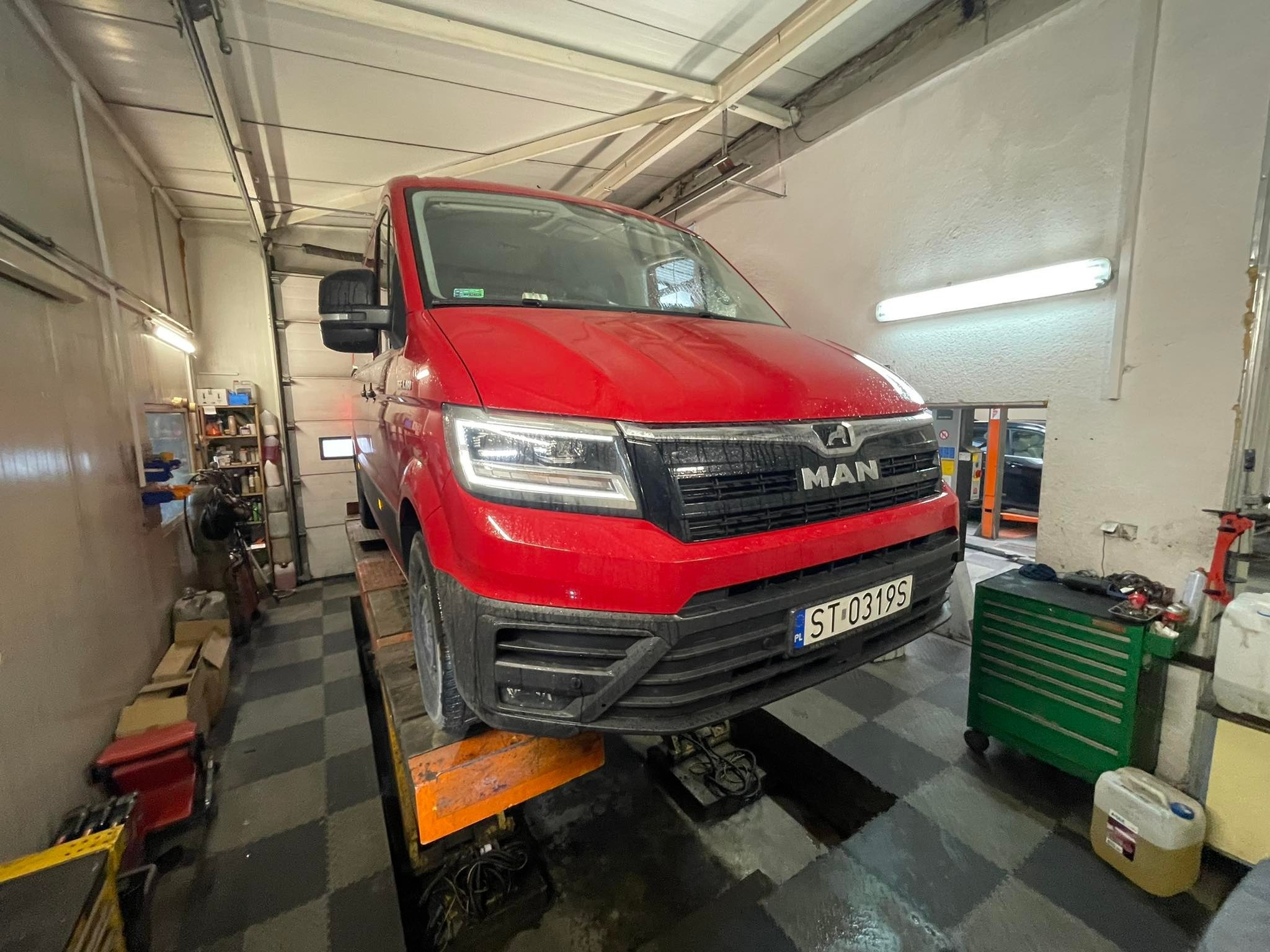 Czerwony samochód dostawczy MAN na podnośniku w warsztacie, widoczny numer rejestracyjny ST-0319S, obok zielony wózek narzędziowy.