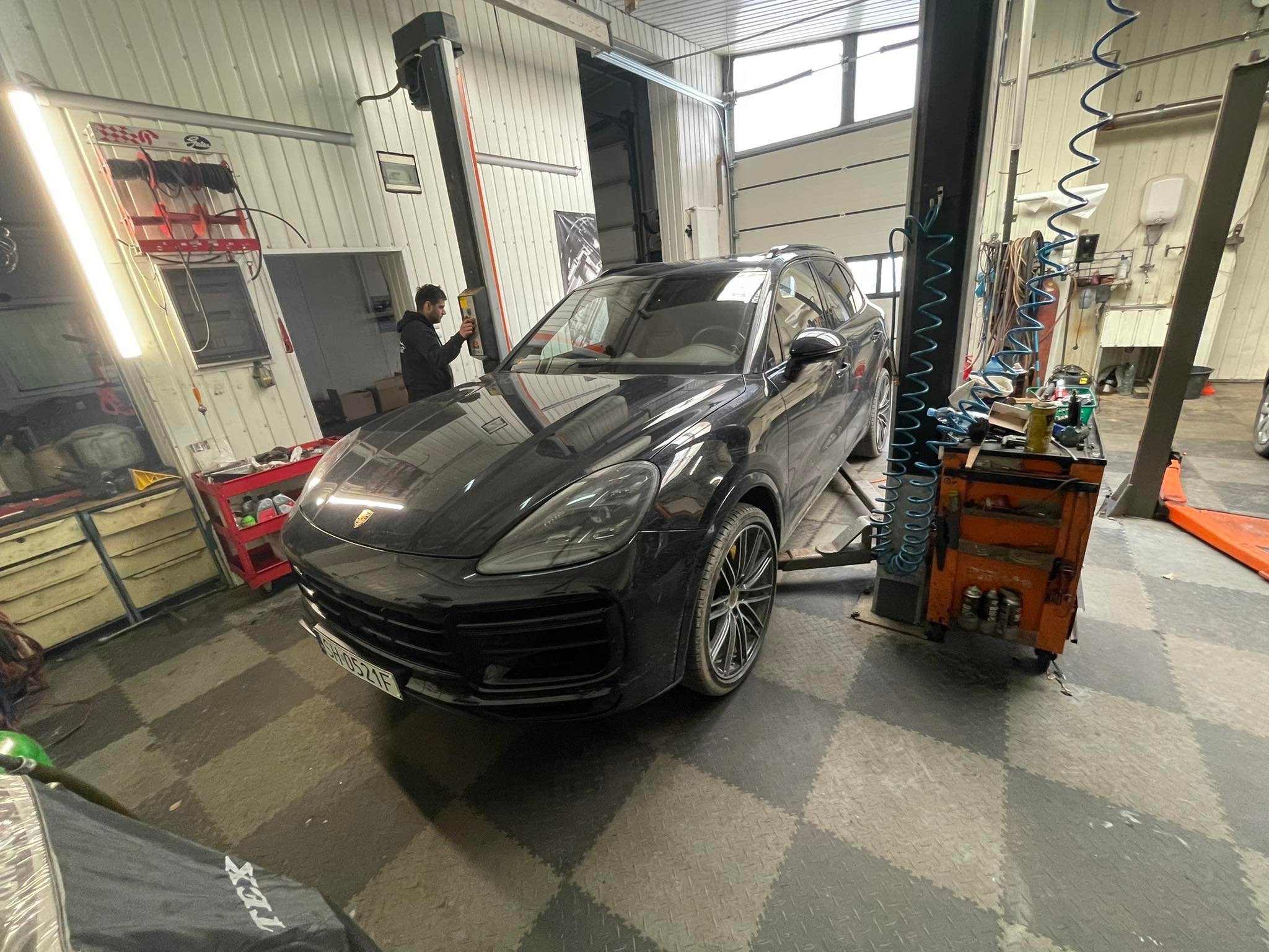 Czarny SUV na podnośniku w warsztacie samochodowym, widoczny pracownik obsługujący panel sterowania podnośnikiem, na podłodze szaro-czarna szachownica z gumy.