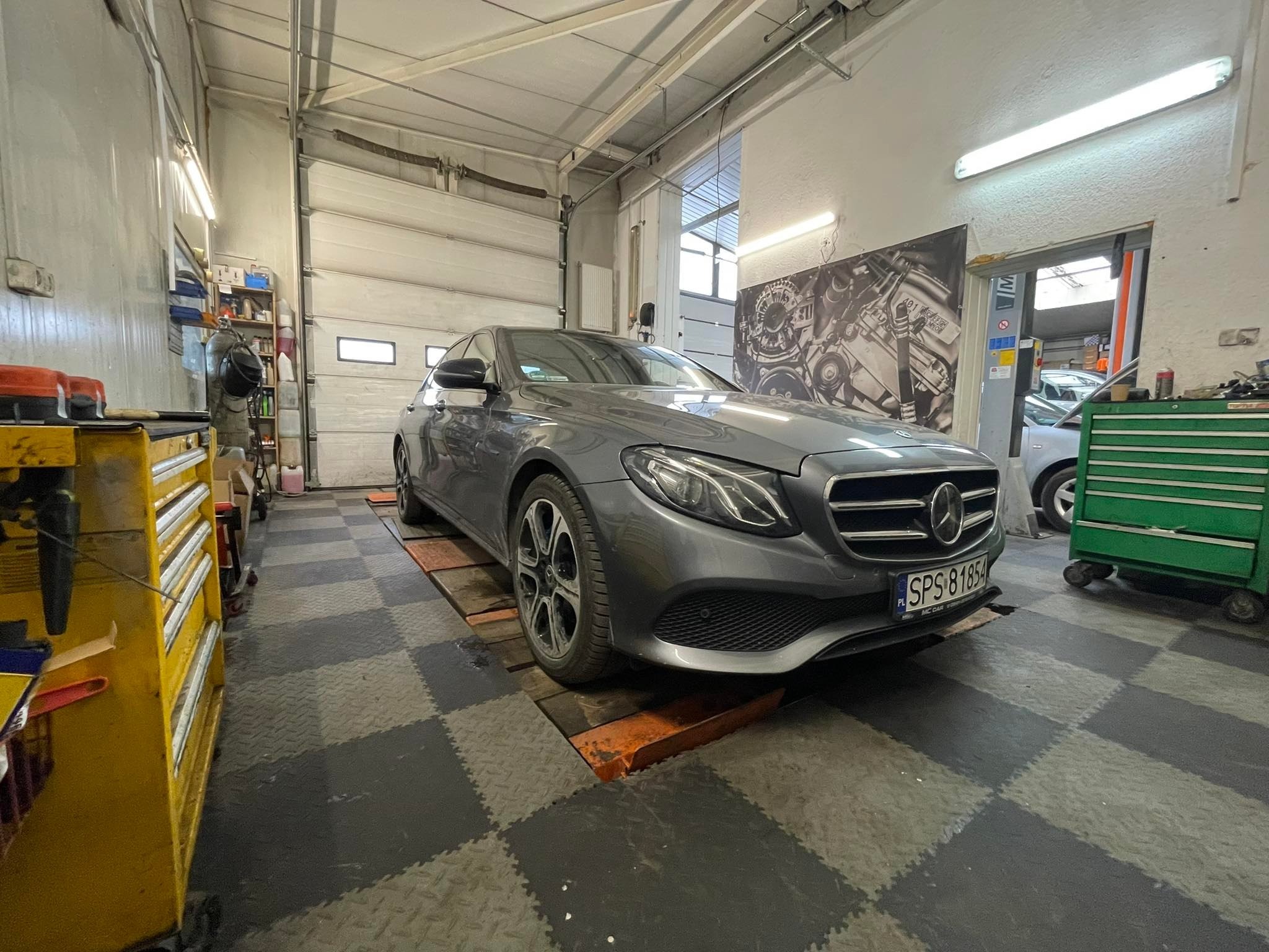Szary Mercedes-Benz E-Klasa na stanowisku warsztatowym z podnośnikiem, widoczny żółty wózek narzędziowy i plakat z silnikiem na ścianie.