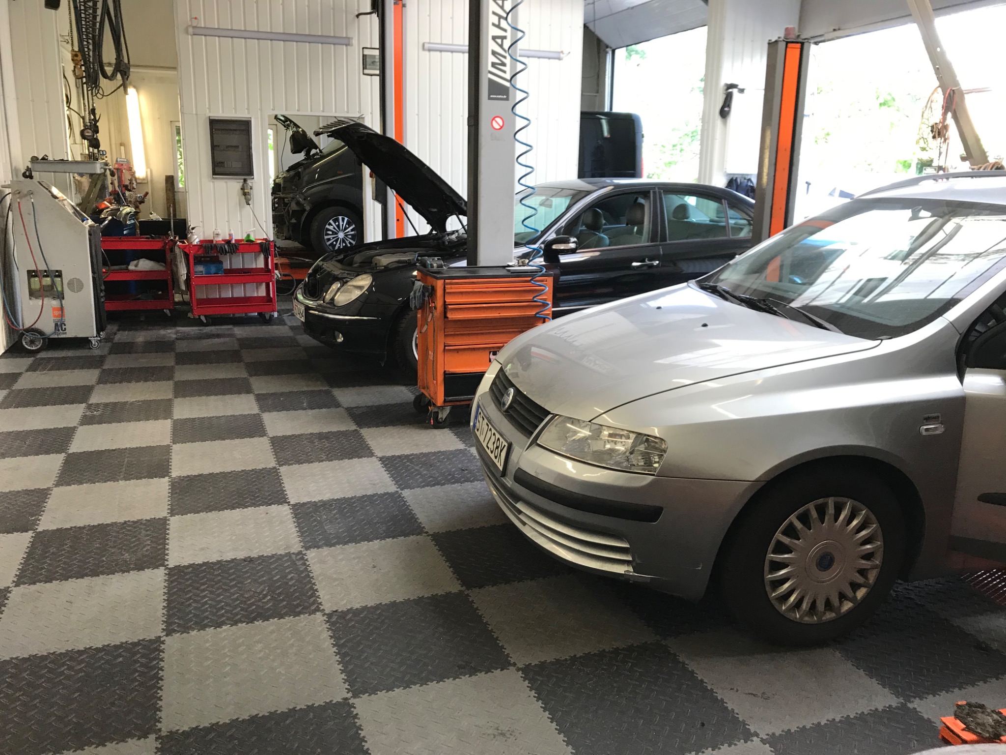 Wnętrze warsztatu samochodowego z trzema samochodami: srebrny Fiat Stilo na pierwszym planie, czarny Mercedes z otwartą maską na podnośniku i czarny samochód w tle. Widoczne narzędzia warsztatowe...