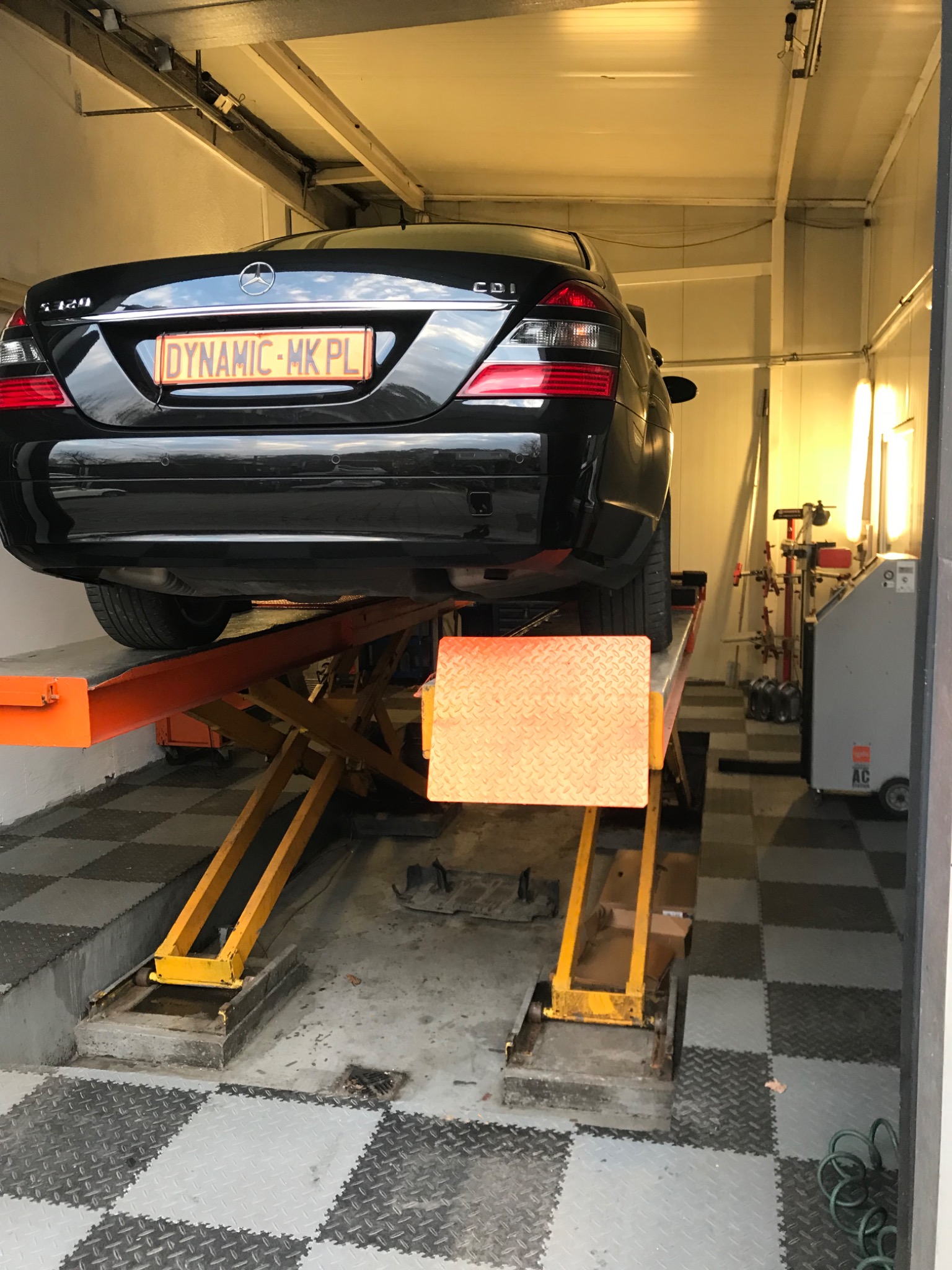 Czarny Mercedes klasy S na podnośniku hydraulicznym w warsztacie, widok z tyłu, tablica rejestracyjna 'DYNAMIC-MK.PL'.