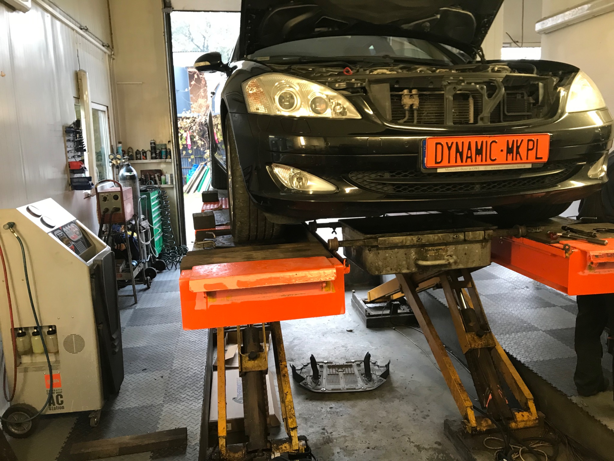 Czarny samochód z otwartą maską uniesiony na podnośniku w warsztacie, widoczna pomarańczowa tablica rejestracyjna z napisem DYNAMIC MK PL, obok stoi maszyna do obsługi klimatyzacji.
