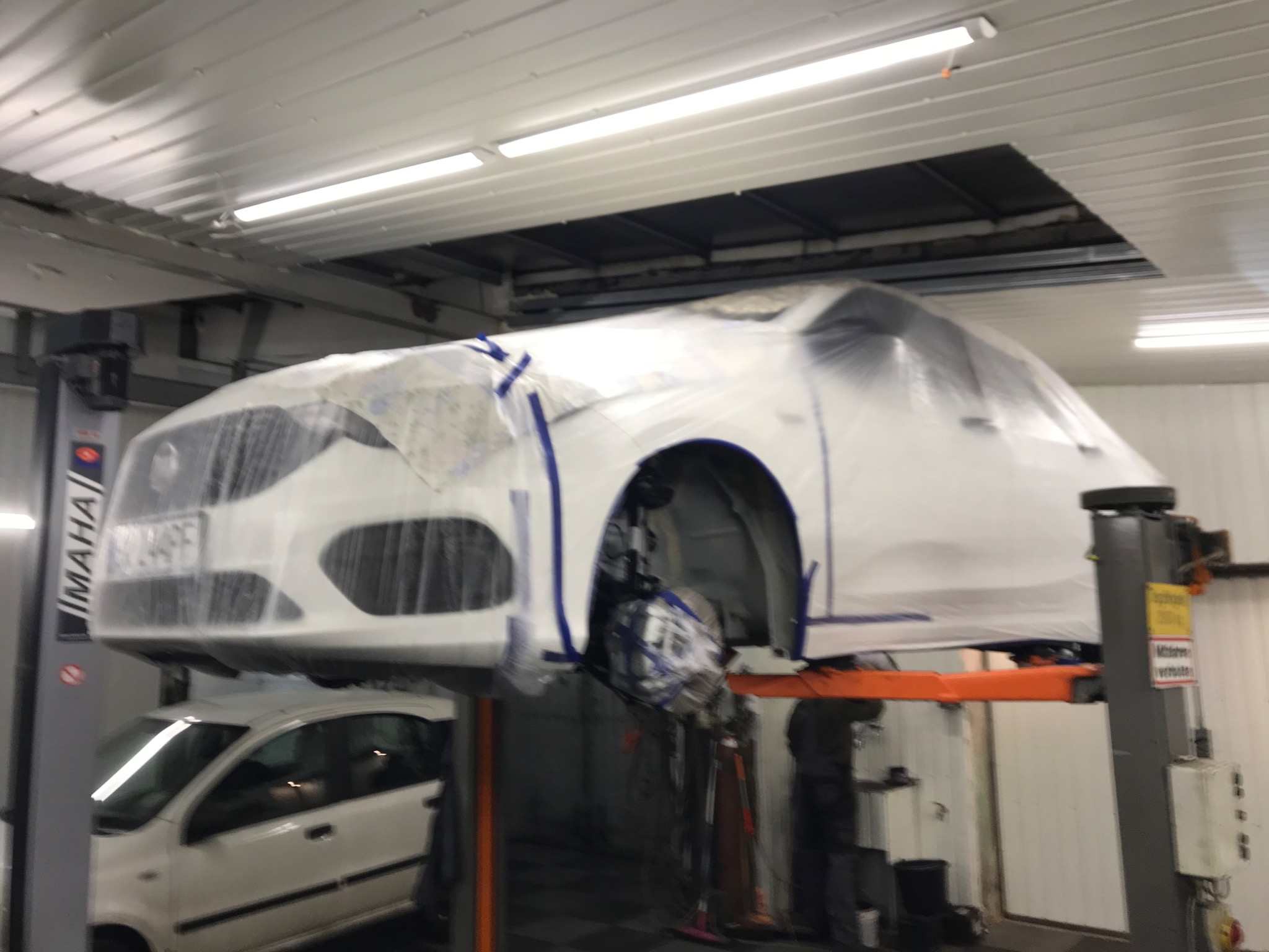 Biały samochód osobowy na podnośniku w warsztacie, zabezpieczony folią i taśmą malarską przed pracami lakierniczymi lub konserwacją antykorozyjną. Widoczna dolna część innego samochodu pod spodem.