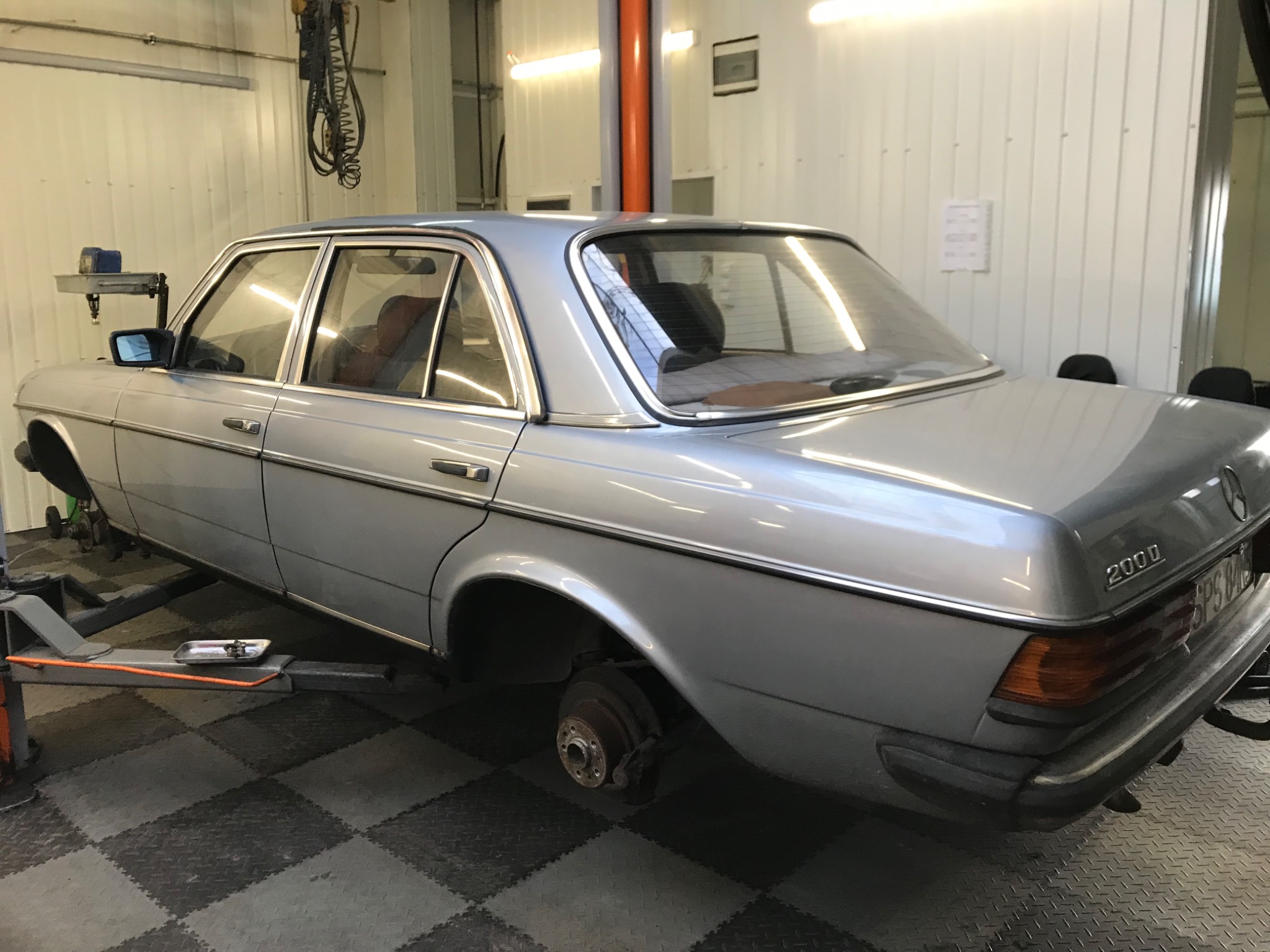 Srebrny Mercedes 200D na podnośniku w warsztacie, bez jednego koła, widoczna tarcza hamulcowa i elementy zawieszenia.