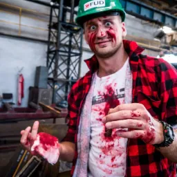 Mężczyzna w zielonym kasku FUD i czerwonej koszuli w kratę, z teatralną krwią na twarzy i ubraniu, demonstruje opatrywanie rany podczas pozorowanego wypadku na tle hali produkcyjnej.