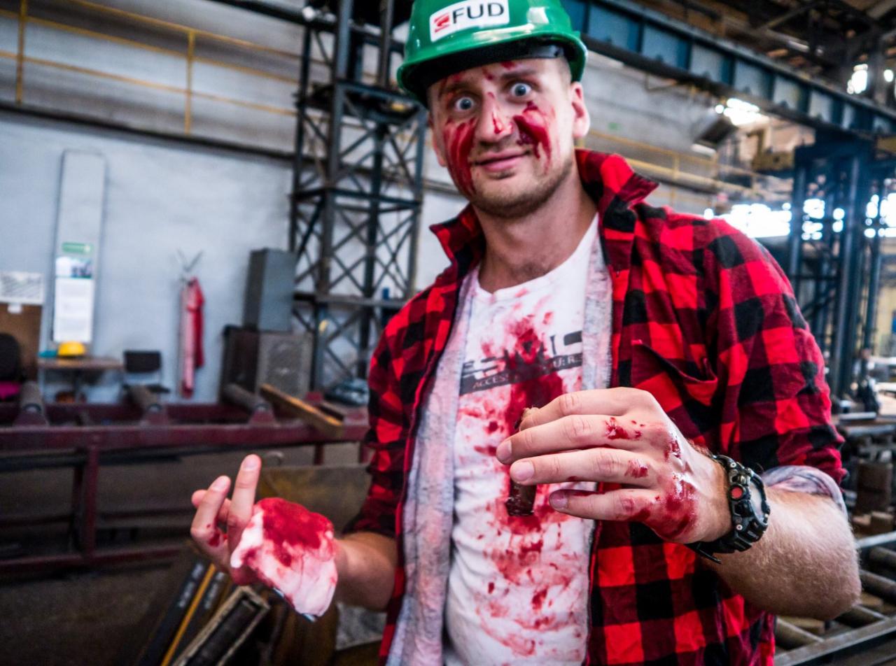 Mężczyzna w zielonym kasku FUD i czerwonej koszuli w kratę, z teatralną krwią na twarzy i ubraniu, demonstruje opatrywanie rany podczas pozorowanego wypadku na tle hali produkcyjnej.