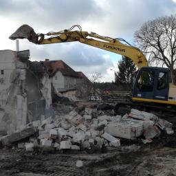 Wilkam - Żółto-niebieska koparka Furukawa 730 kruszy resztki betonowej konstrukcji z graffiti na tle zabudowań mieszkalnych i drzewa bez liści.