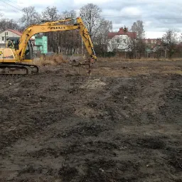 Żółta koparka Furukawa na placu budowy, świeżo przekopana ziemia, w tle zabudowa jednorodzinna, pochmurne niebo.