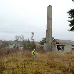 Wilkam - Pracownik w kamizelce odblaskowej idzie przez zarośnięte pole w kierunku częściowo zburzonego budynku z wysokim kominem i panelami słonecznymi na dachu.