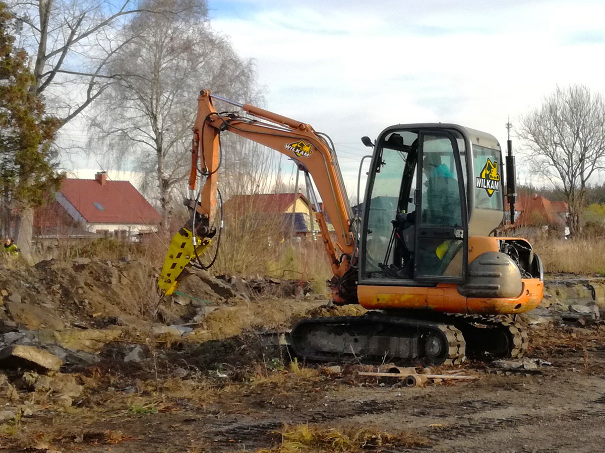 Pomarańczowa mini-koparka z hydraulicznym młotem marki Wilkam rozbija betonowe elementy na placu budowy, w tle widoczne domy jednorodzinne i drzewa bezlistne.