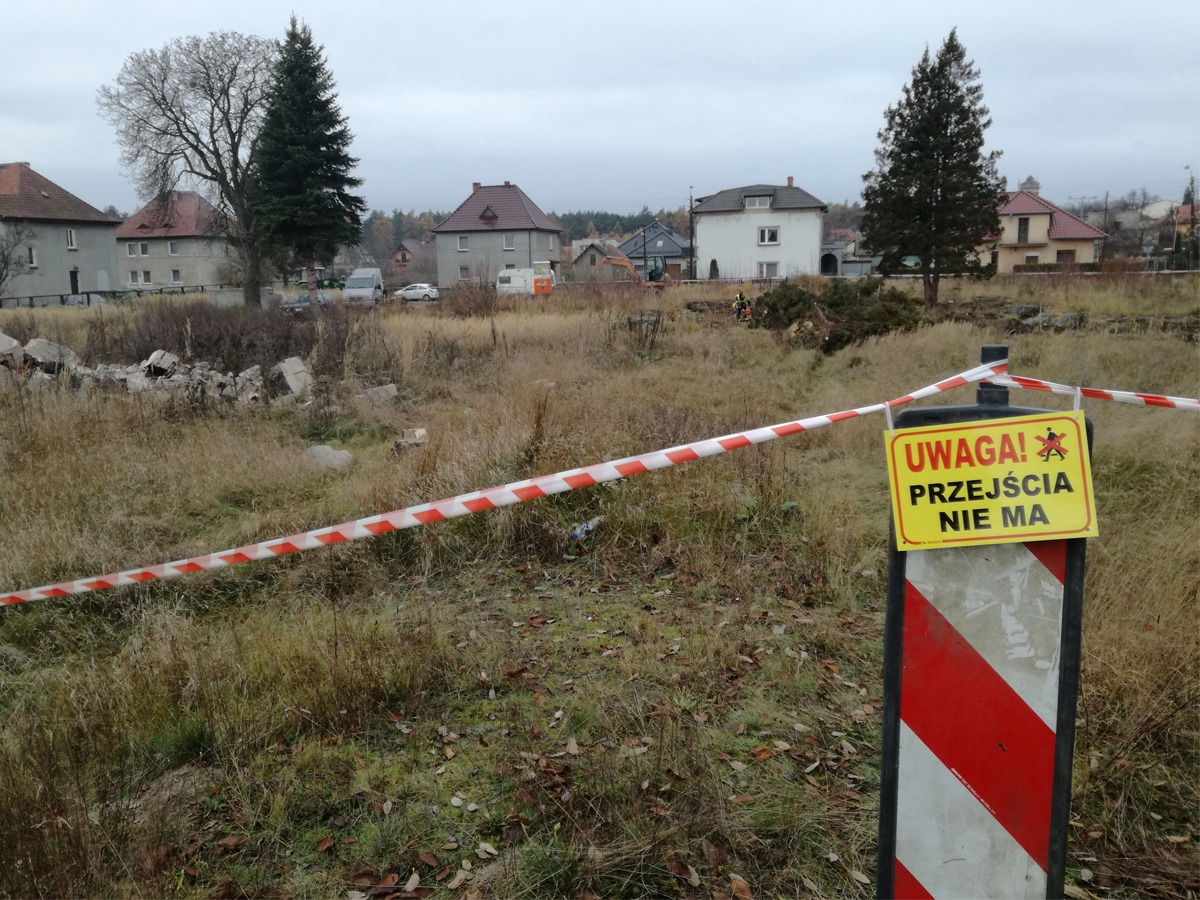 Teren po wyburzeniu z gruzem, zabezpieczony taśmą ostrzegawczą i znakiem 'Uwaga! Przejścia nie ma', w tle budynki mieszkalne i drzewa.