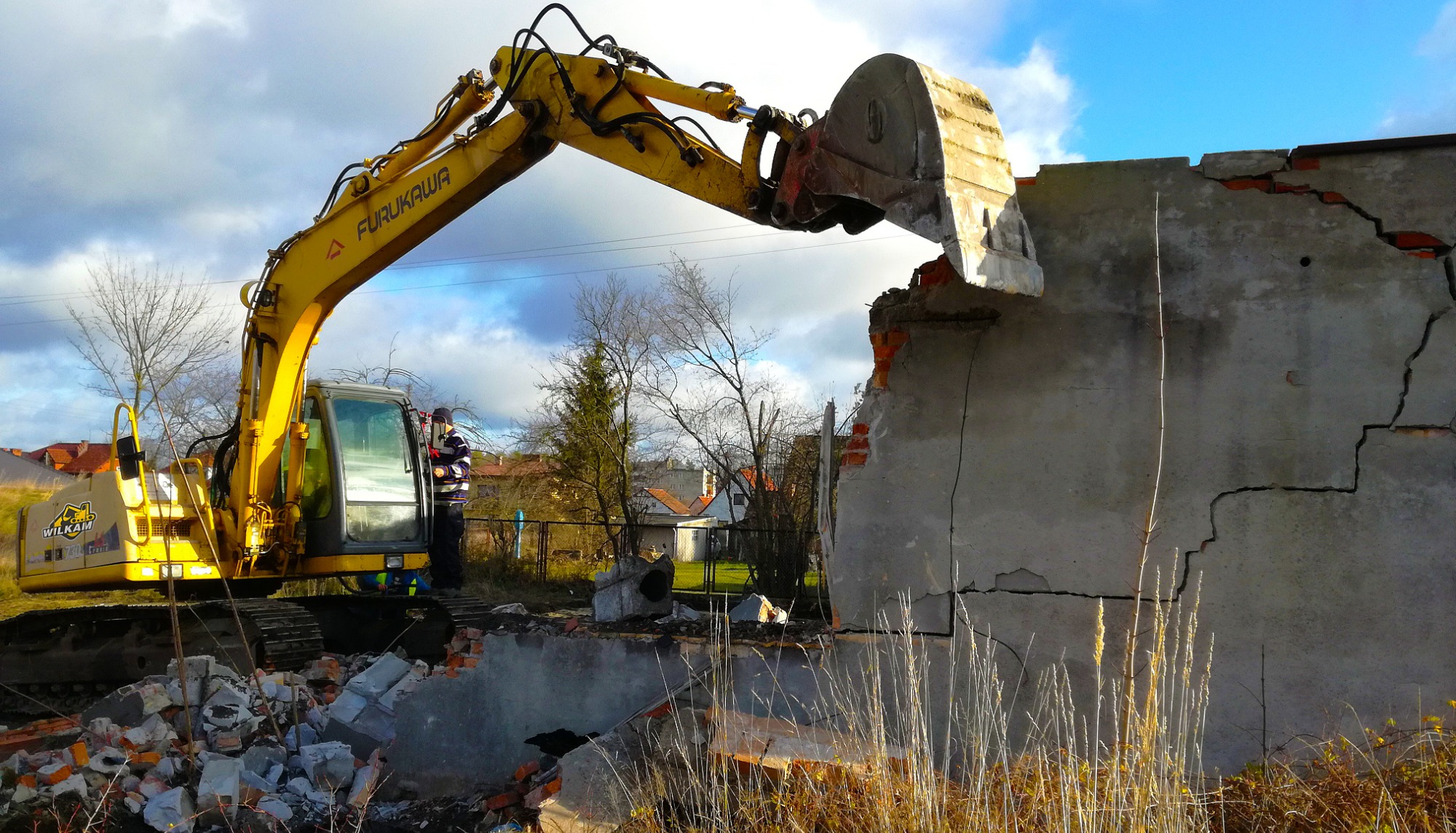 Żółta koparka Furukawa z operatorem w kabinie kruszy betonową ścianę budynku z widocznymi pęknięciami, na tle błękitnego nieba z chmurami i zabudowań mieszkalnych.