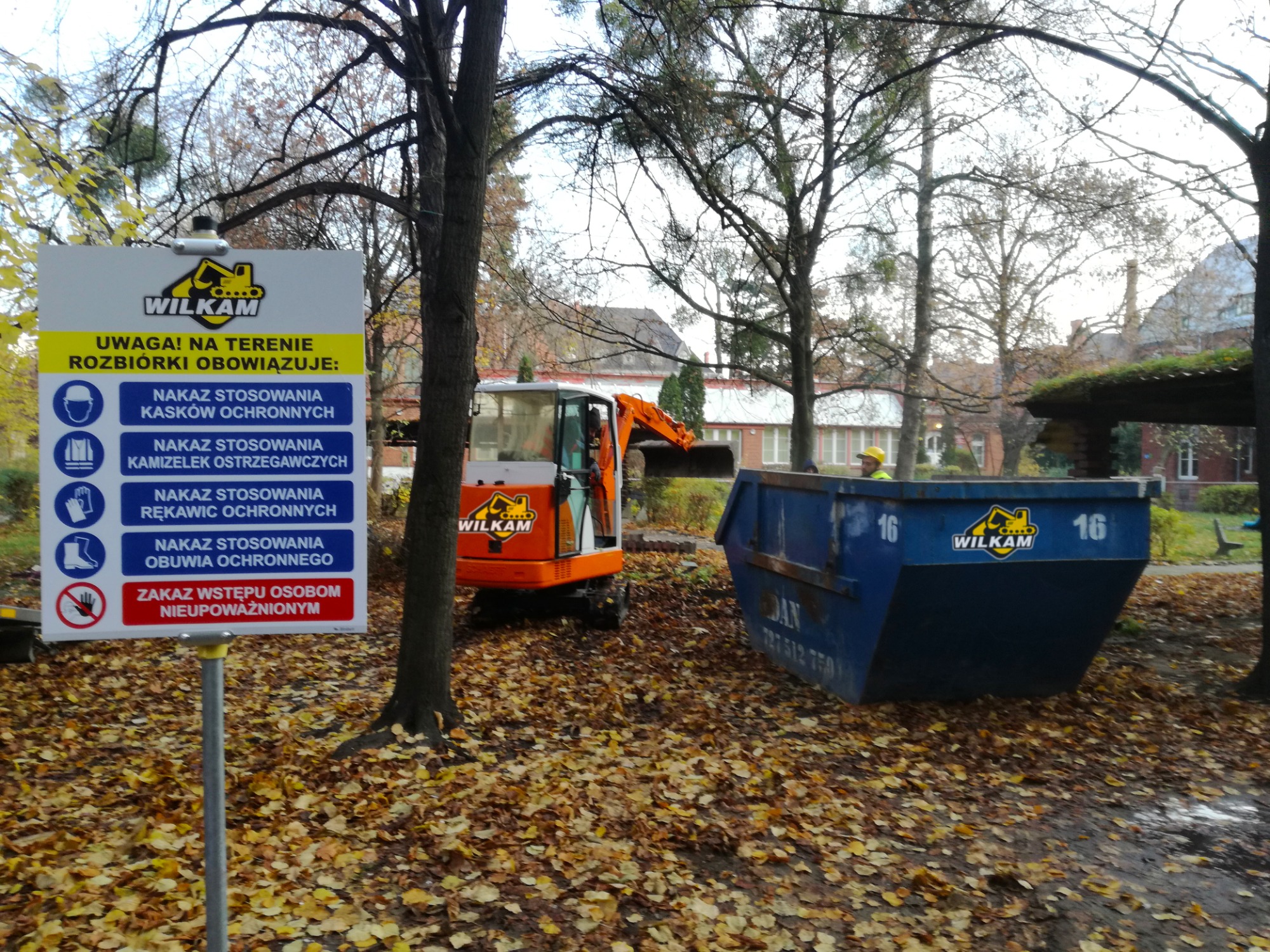 Pomarańczowa minikoparka na placu budowy w jesiennej scenerii, obok kontener na gruz i tablica informacyjna z nakazem stosowania środków ochrony osobistej.