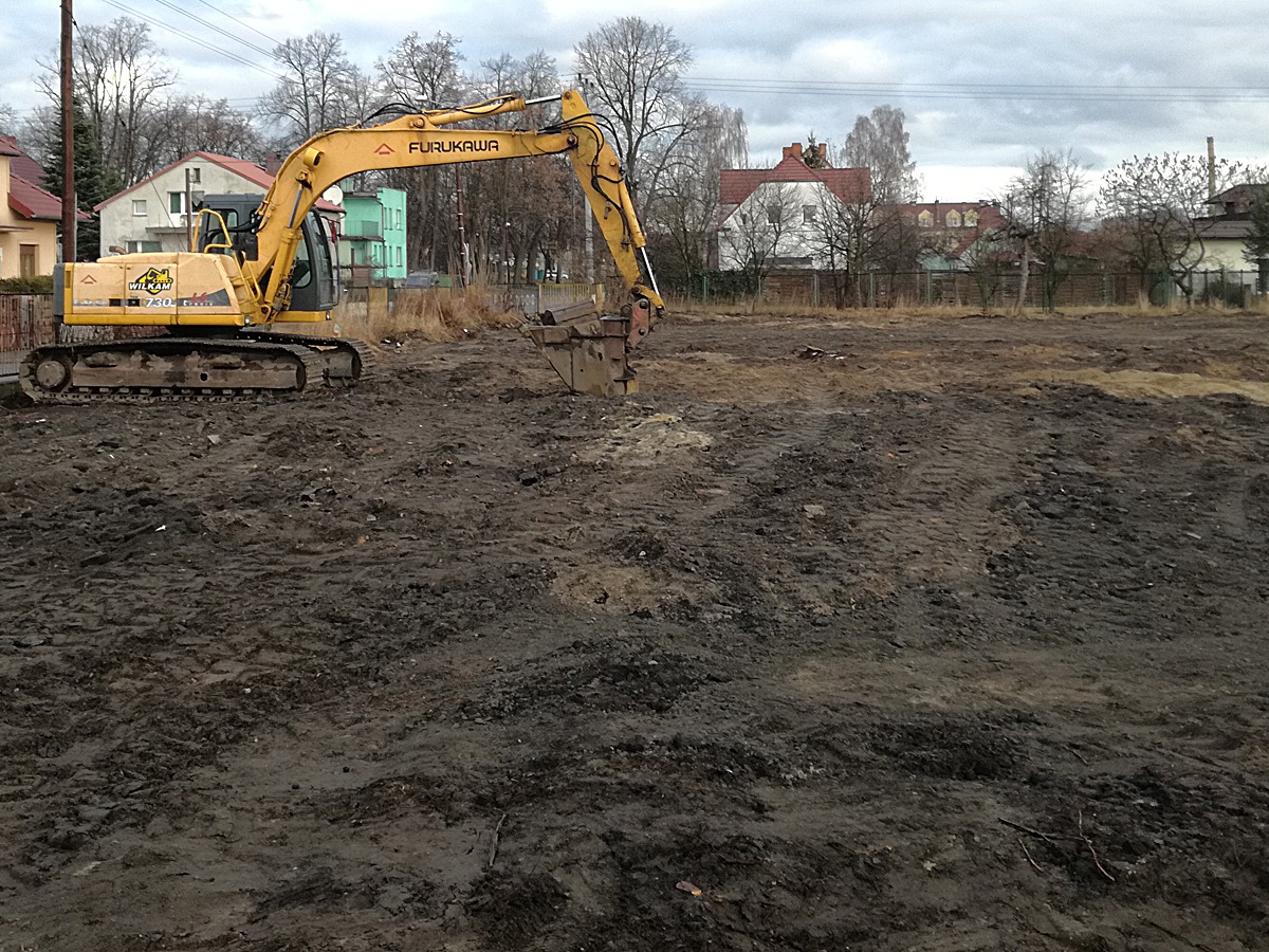 Żółta koparka Furukawa na placu budowy, świeżo przekopana ziemia, w tle zabudowa jednorodzinna, pochmurne niebo.